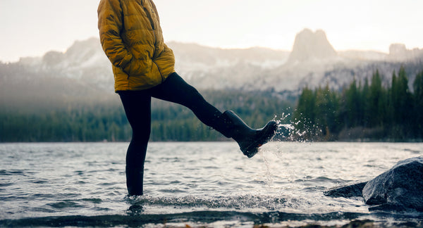 Person in yellow jacket and dark pants stepping into water with mountains and trees in the background